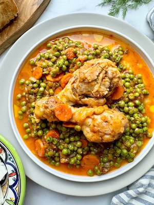 Chicken with green peas served on white plate with slices of bread in the background.