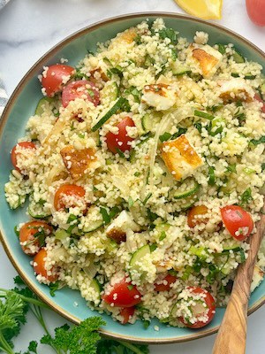 Halloumi and couscous salad served on a plate next to lemon slices and cherry tomatoes.