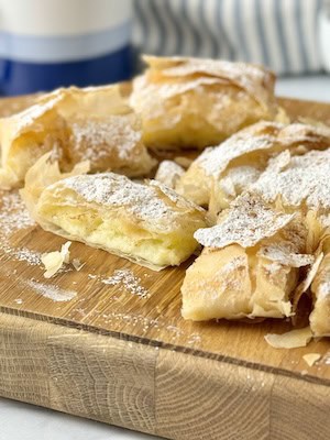 A bougatsa parcel cut into squares on a wooden board with powdered sugar and cinnamon on top.