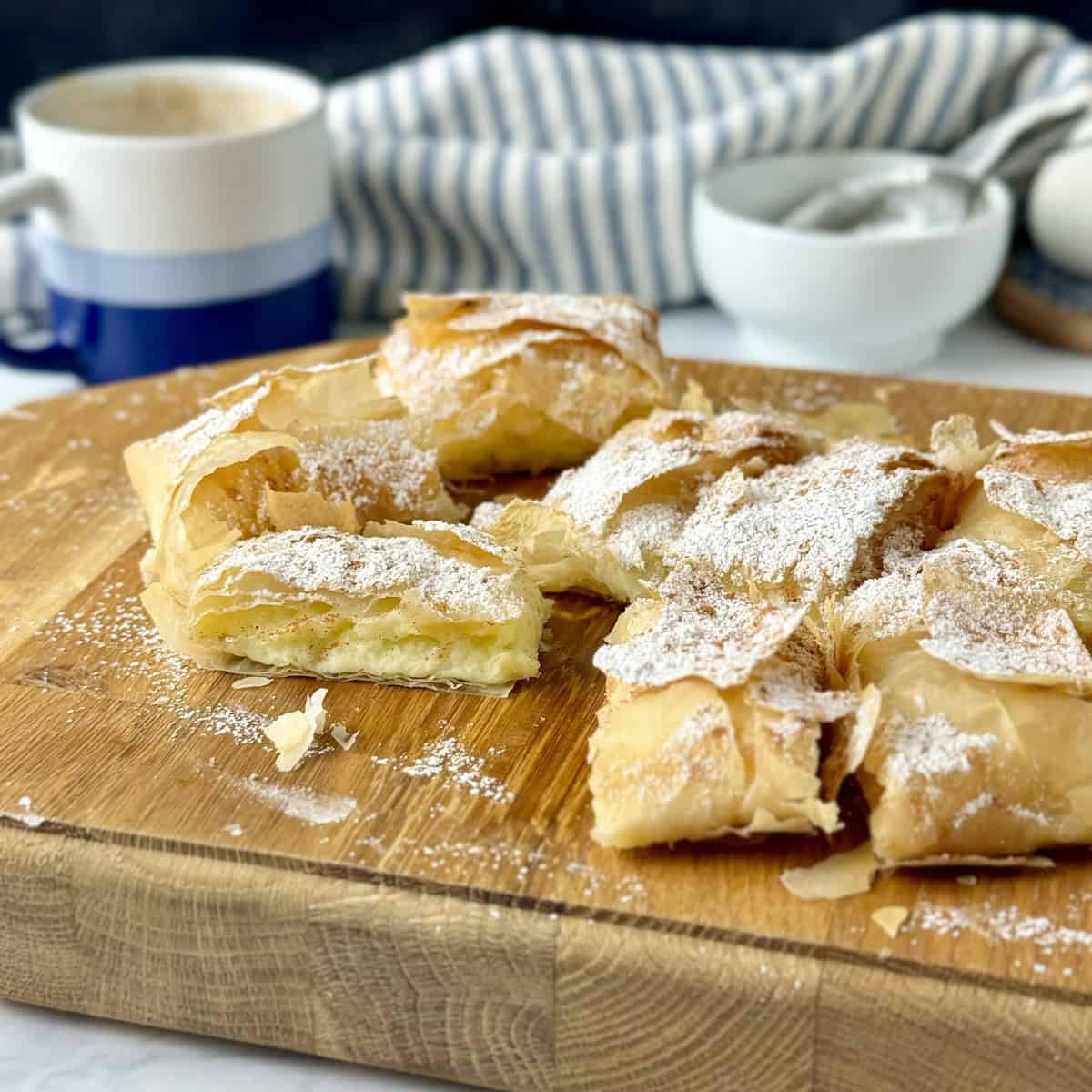 A bougatsa parcel cut into squares on a wooden board with powdered sugar and cinnamon on top.