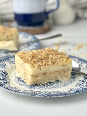 A slice of cream and biscuit cake served on blue porcelain dish with a mug of coffee and flowers in the background.