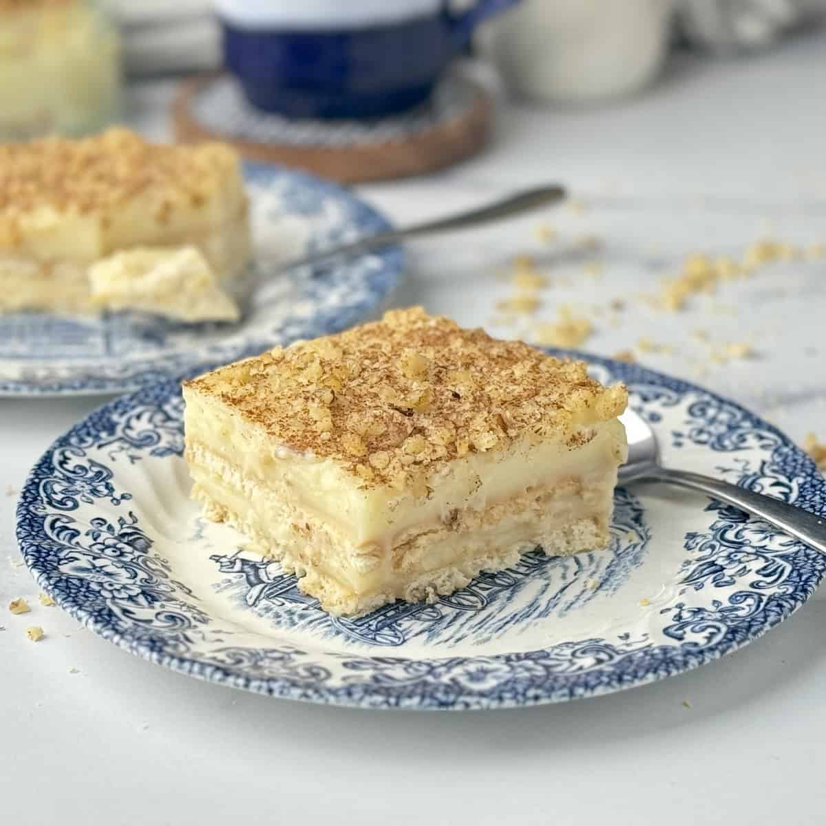 A slice of cream and biscuit cake served on blue porcelain dish with a mug of coffee and flowers in the background.