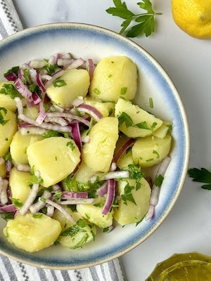 Potato salad served in a bowl next to olive oil bottle, lemon and fresh parsley.