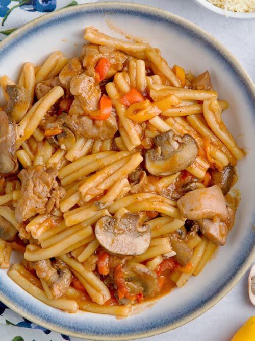 Chicken and mushroom pasta in a white and blue bowl next to raw mushrooms and peppers and bowl with parmesan cheese.