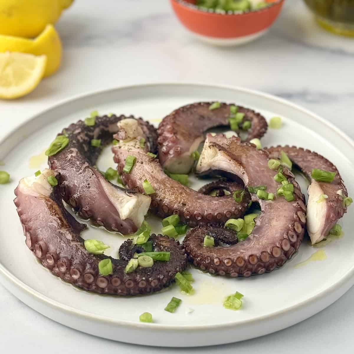 Pan-seared octopus with chopped green onions on a white plate next to lemons and olive oil.