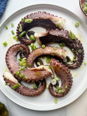 Pan-seared octopus with chopped green onions on a white plate next to lemons and olive oil.